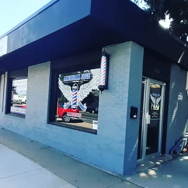 American Edge Barbershop storefront: blue-grey building with the winged barber-pole logo in the window and a barber pole by the door